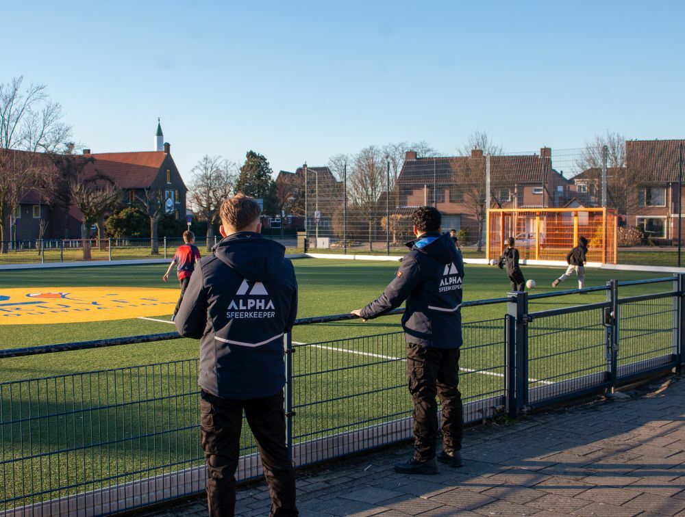 Twee straatcoaches staan langs een voetbalveld met spelende kinderen.